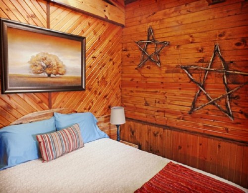 Cozy bedroom with wooden walls, a framed painting, and rustic star decorations. A bed with blue pillows and a striped throw.