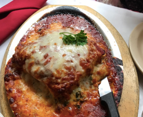 A sizzling plate of lasagna topped with melted cheese and marinara sauce, garnished with a sprig of parsley.