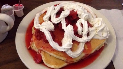 A stack of pancakes topped with strawberry sauce and whipped cream, served on a white plate.