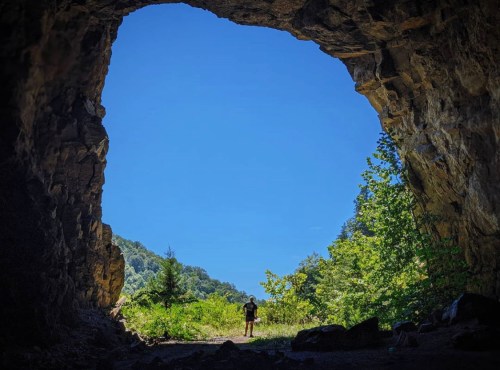 A person stands at the entrance of a cave, framed by rocky walls and surrounded by lush greenery and a clear blue sky.