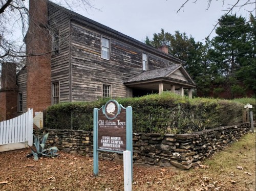 Historic wooden house with a stone wall and sign for Old Alabama Town, surrounded by trees and grass.