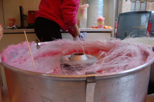 A person in a pink sweater prepares cotton candy in a metal machine, with fluffy pink strands forming around it.