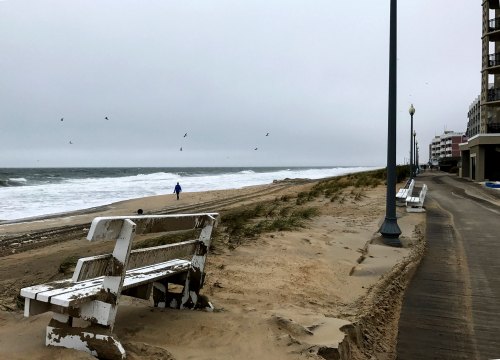 A cloudy beach scene with a person walking along the shore, benches on the sand, and waves crashing in the background.