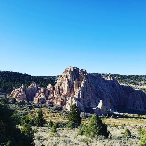 Rocky formations rise against a clear blue sky, surrounded by green trees and open land.