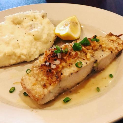 A plate featuring a piece of fish topped with herbs, served with creamy mashed potatoes and a lemon wedge.