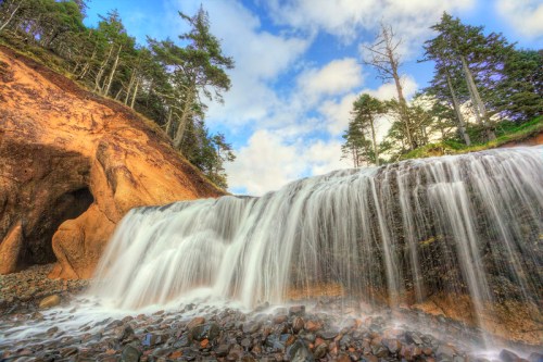 A serene waterfall cascades over rocks, surrounded by trees and a blue sky with fluffy clouds.