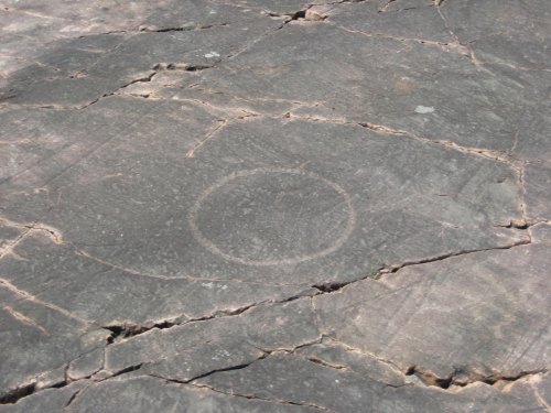 A circular petroglyph etched into a rocky surface, surrounded by cracks and natural textures.