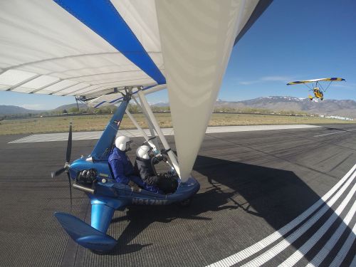 A blue ultralight aircraft on a runway with two pilots inside, while another ultralight flies overhead against a clear sky.