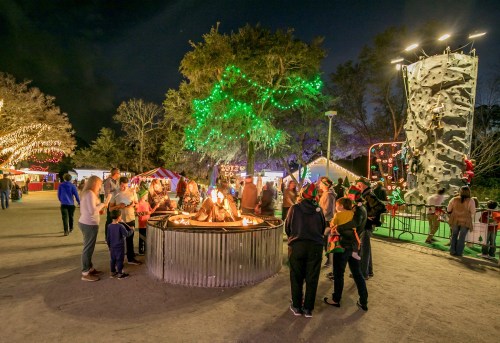 A festive outdoor scene with people gathered around a fire pit, colorful lights, and a rock climbing wall in the background.