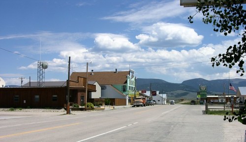 A small town street with buildings, a radio tower, and mountains under a blue sky with fluffy clouds.