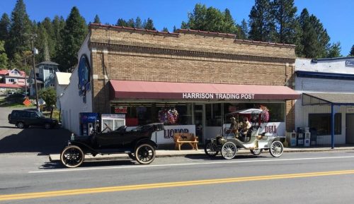 Two vintage cars parked in front of the Harrison Trading Post, a brick building with a sign and colorful decorations.