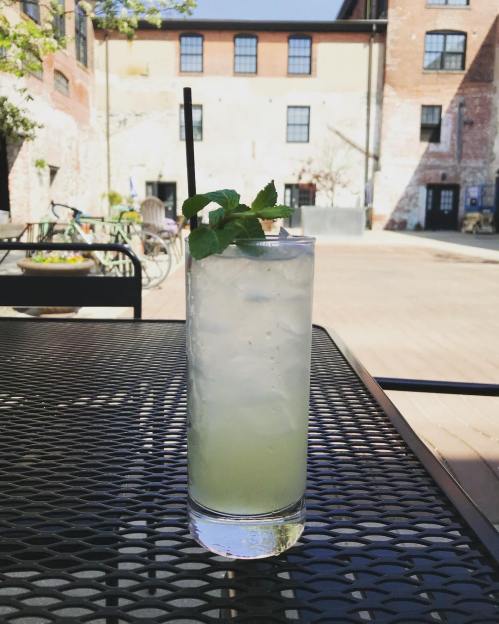 A refreshing cocktail with mint leaves sits on a black metal table in a sunny courtyard.