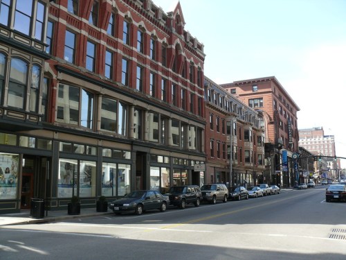 A city street lined with historic brick buildings and parked cars under a clear blue sky.