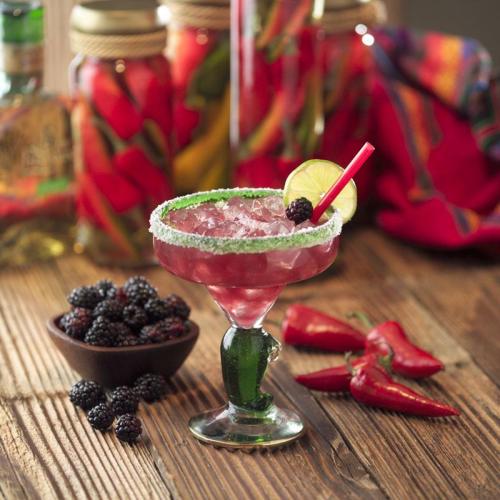 A vibrant cocktail with a lime slice and blackberries, served in a decorative glass, surrounded by peppers and berries.