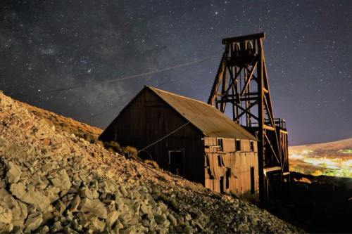 An old mining structure under a starry night sky, surrounded by rocky terrain and distant city lights.