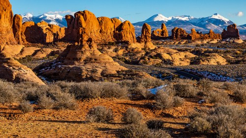 A desert landscape featuring red rock formations and snow-capped mountains in the background under a clear blue sky.