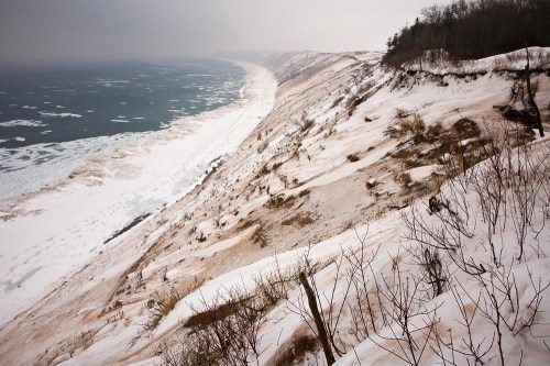 A snowy cliffside overlooking a frozen lake, with ice patches and a misty horizon in the background.