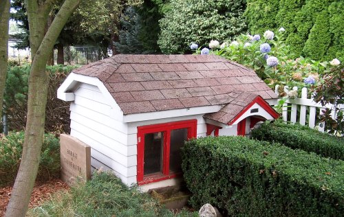 A small, charming white playhouse with a red roof and trim, surrounded by greenery and flowers.