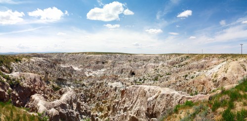 A panoramic view of a rugged, colorful canyon landscape under a bright blue sky with scattered clouds.