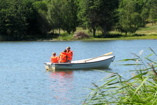 Three people in bright orange life jackets fishing from a small boat on a calm lake surrounded by greenery.