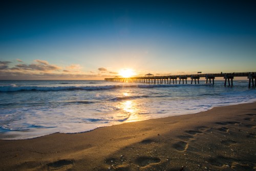A serene beach scene at sunset, with a pier extending into the ocean and gentle waves lapping at the shore.