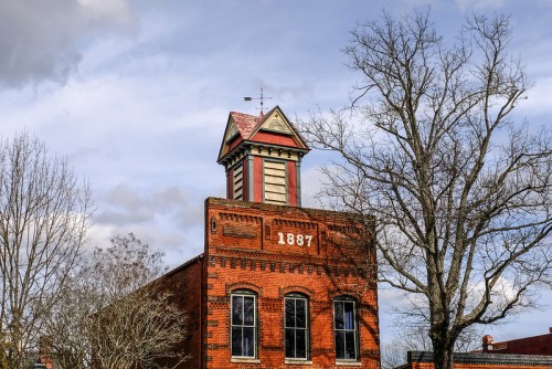 Historic red brick building with a tower, marked "1887," surrounded by bare trees under a cloudy sky.