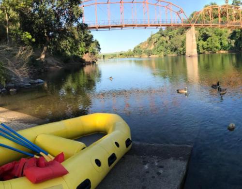 A yellow raft on the shore of a calm river, with ducks swimming and a red bridge in the background.