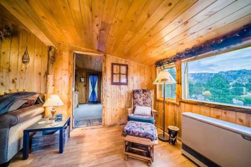 Cozy wooden interior of a cabin with a chair, lamp, and large window showcasing mountain views.