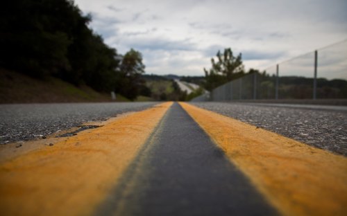 A close-up view of a two-lane road with a yellow center line, surrounded by trees and a cloudy sky.