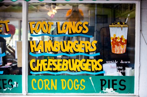 Colorful window sign displaying menu items: Foot Longs, Hamburgers, Cheeseburgers, Corn Dogs, and Pies.