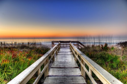 A wooden boardwalk leads to a serene beach at sunset, surrounded by lush greenery and calm waters.