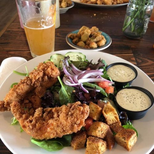 A plate of fried chicken on a bed of mixed greens, tomatoes, and croutons, with two dipping sauces and a beer.