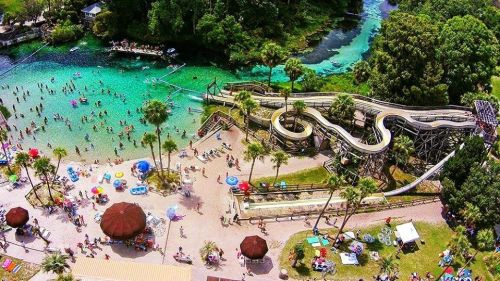 Aerial view of a water park with slides, pools, and people enjoying the sunny day surrounded by lush greenery.