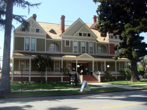 A large, colorful Victorian-style house with a manicured lawn and trees in front, featuring multiple gables and porches.