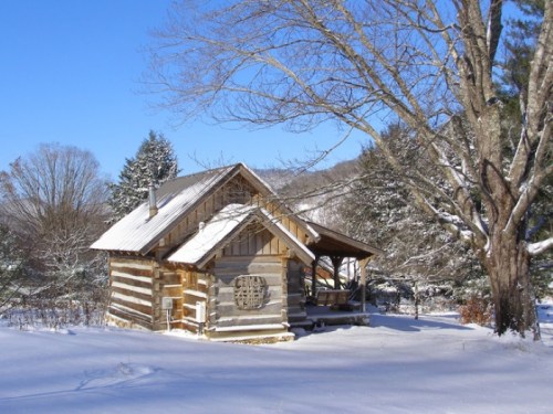 A cozy log cabin surrounded by snow-covered trees under a clear blue sky.