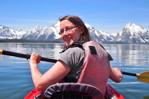 A person in a kayak smiles, surrounded by mountains and calm water on a sunny day.