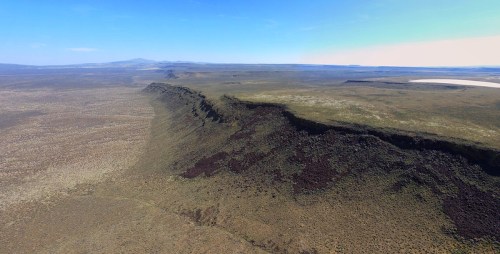 Aerial view of a vast, flat landscape with a prominent, rugged cliff edge under a clear blue sky.