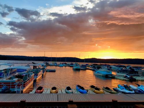 Sunset over a marina with boats docked, reflecting vibrant colors on the water and a cloudy sky.