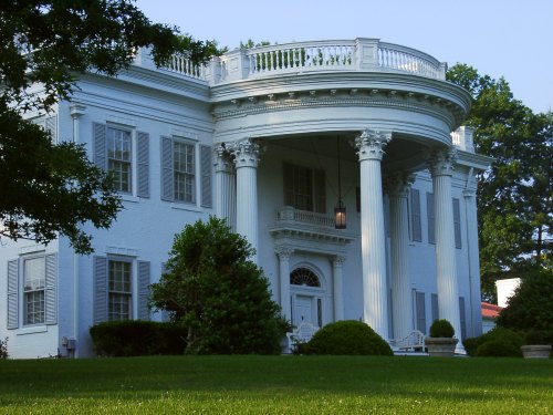 A grand white mansion with a circular porch, tall columns, and lush green landscaping.