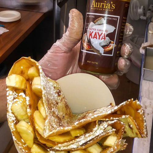 A hand holds a bottle of salted caramel kaya next to a plate of banana-filled crepes dusted with powdered sugar.
