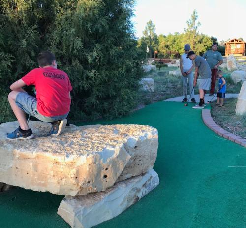 A group of children and adults playing mini-golf outdoors, with one child sitting on a rock.