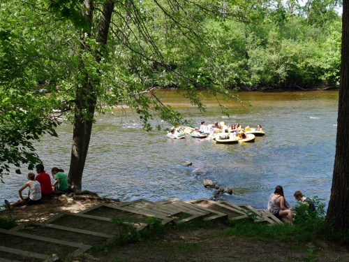 A group of people in colorful rafts float on a river, while others relax on the shore under leafy trees.