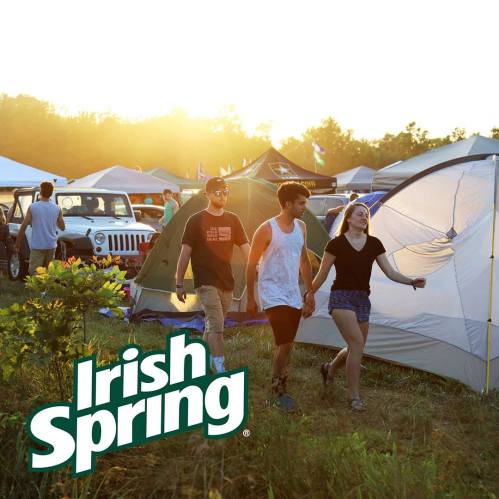 Three friends walk through a campsite at sunset, surrounded by tents and vehicles, with a vibrant outdoor atmosphere.