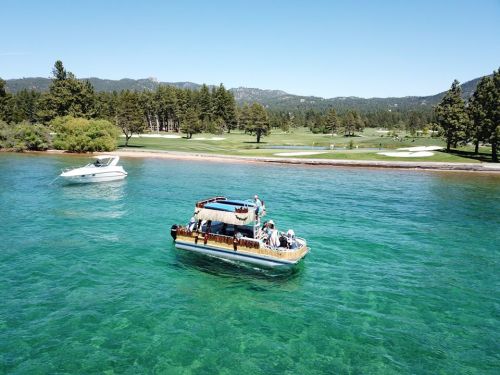 A boat with people floats on clear blue water near a green shoreline and trees under a bright blue sky.