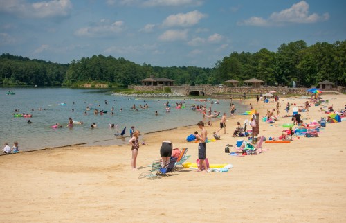 A sunny beach scene with people swimming, sunbathing, and relaxing on colorful towels and chairs by the water.