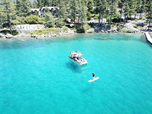 Aerial view of a clear turquoise lake with a boat and a person paddleboarding near the shore. Pine trees in the background.