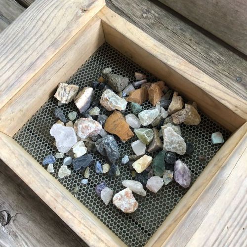 A wooden box containing a variety of colorful gemstones and minerals on a mesh bottom.