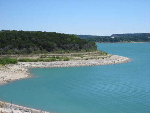 A serene lake with turquoise water, surrounded by green trees and a rocky shoreline under a clear blue sky.