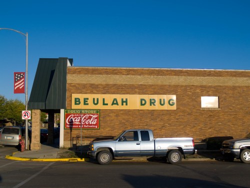 Exterior of Beulah Drug store with a blue pickup parked outside and a Coca-Cola sign visible. Clear blue sky above.
