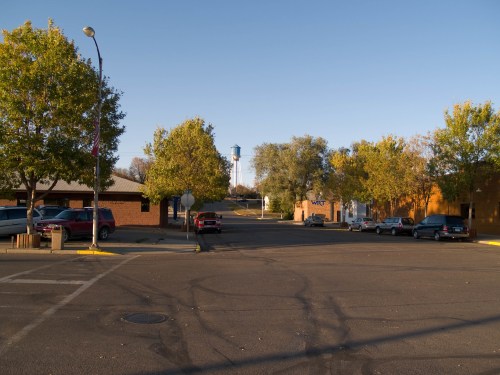 A quiet street scene with trees, parked cars, and a water tower in the distance under a clear blue sky.